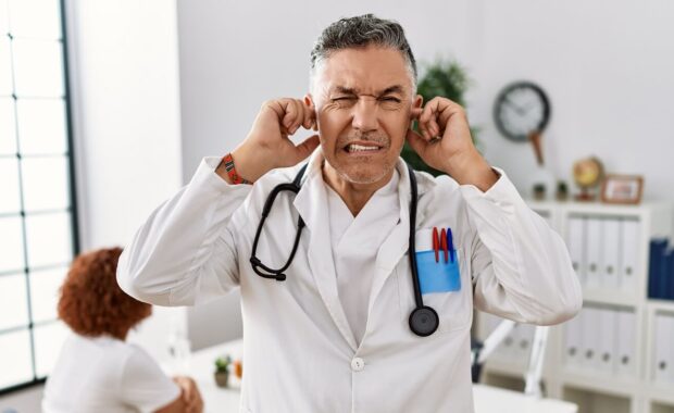 doctor man at the clinic with a patient covering ears with fingers with annoyed expression for the noise of loud music