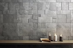 wooden table topped with two candles next to a wall covered in blocks of grey tiles and a candle holder