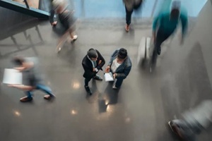 business people standing in the lobby of an office looking at a tablet while people are walking past in a blur
