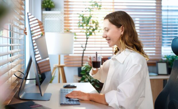 caucasian woman sitting at home office table drinking coffee with desktop pc. Smiling relaxed young lady hybrid working on computer in modern house